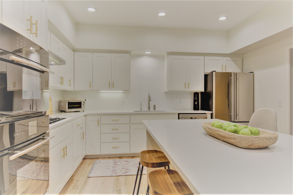 This is an example of a large traditional l-shaped kitchen/diner in Other with a single-bowl sink, shaker cabinets, white cabinets, engineered stone countertops, white splashback, metro tiled splashback, stainless steel appliances, light hardwood flooring, an island and white worktops.