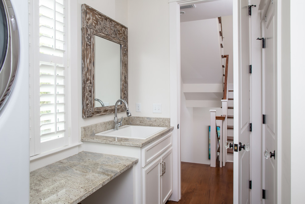 This is an example of a small beach style single-wall laundry room in Charleston with a drop-in sink, white walls, medium hardwood floors, recessed-panel cabinets, white cabinets, granite benchtops and a stacked washer and dryer.