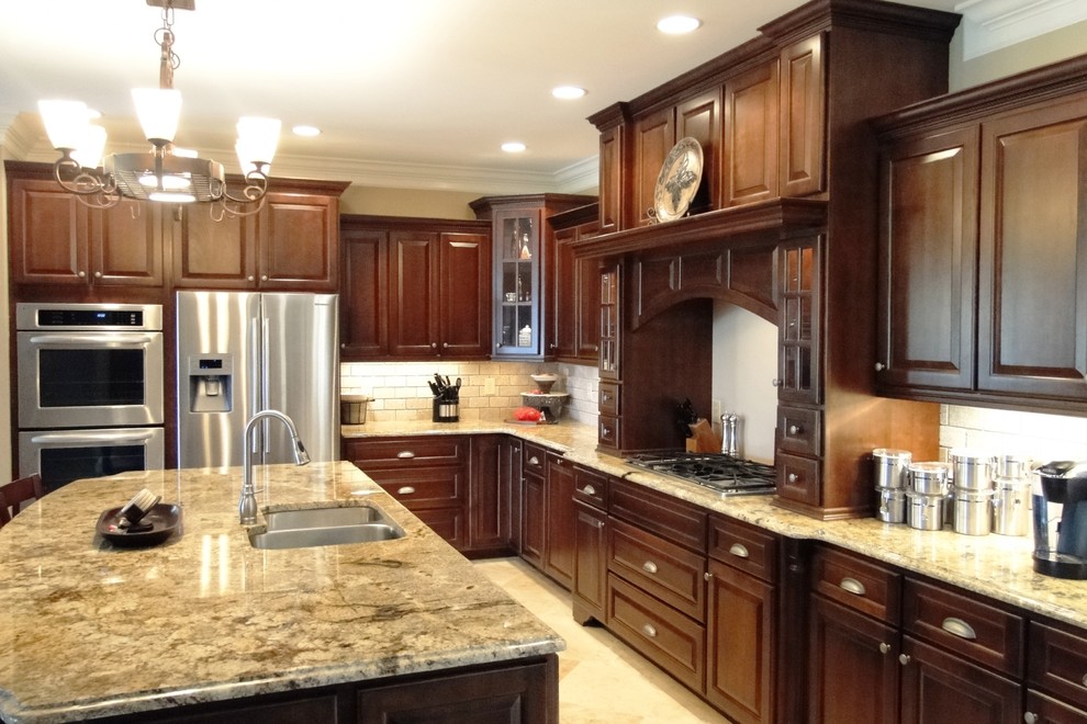 Large transitional l-shaped travertine floor open concept kitchen photo with an undermount sink, raised-panel cabinets, dark wood cabinets, granite countertops, beige backsplash, subway tile backsplash, stainless steel appliances and an island