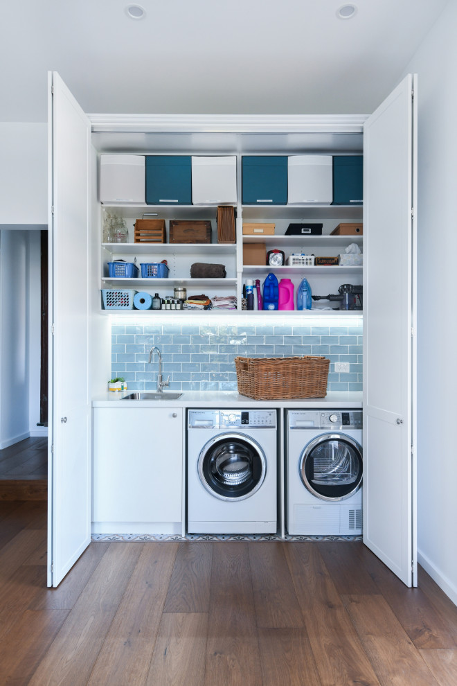 Photo of a contemporary single-wall laundry cupboard in Sydney with an undermount sink, flat-panel cabinets, white cabinets, white walls, dark hardwood floors, a side-by-side washer and dryer, brown floor and white benchtop.