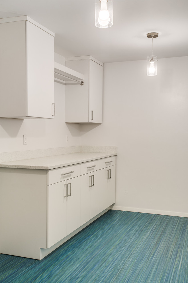 Photo of a mid-sized midcentury l-shaped utility room in Seattle with an utility sink, flat-panel cabinets, white cabinets, white walls, vinyl floors, a stacked washer and dryer and blue floor.