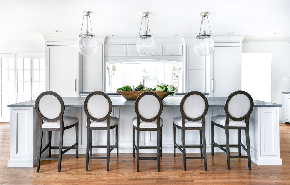Large transitional l-shaped light wood floor eat-in kitchen photo in Philadelphia with a farmhouse sink, flat-panel cabinets, white cabinets, soapstone countertops, white backsplash, ceramic backsplash, paneled appliances, an island and gray countertops