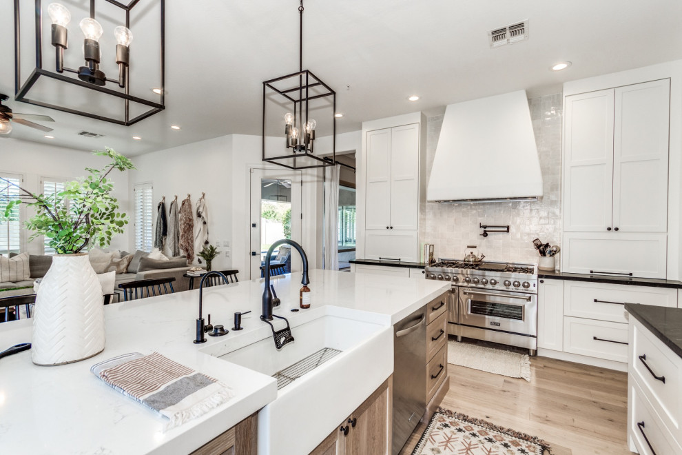This is an example of a large country l-shaped eat-in kitchen in Phoenix with a farmhouse sink, shaker cabinets, light wood cabinets, quartz benchtops, white splashback, terra-cotta splashback, stainless steel appliances, light hardwood floors, with island and white benchtop.