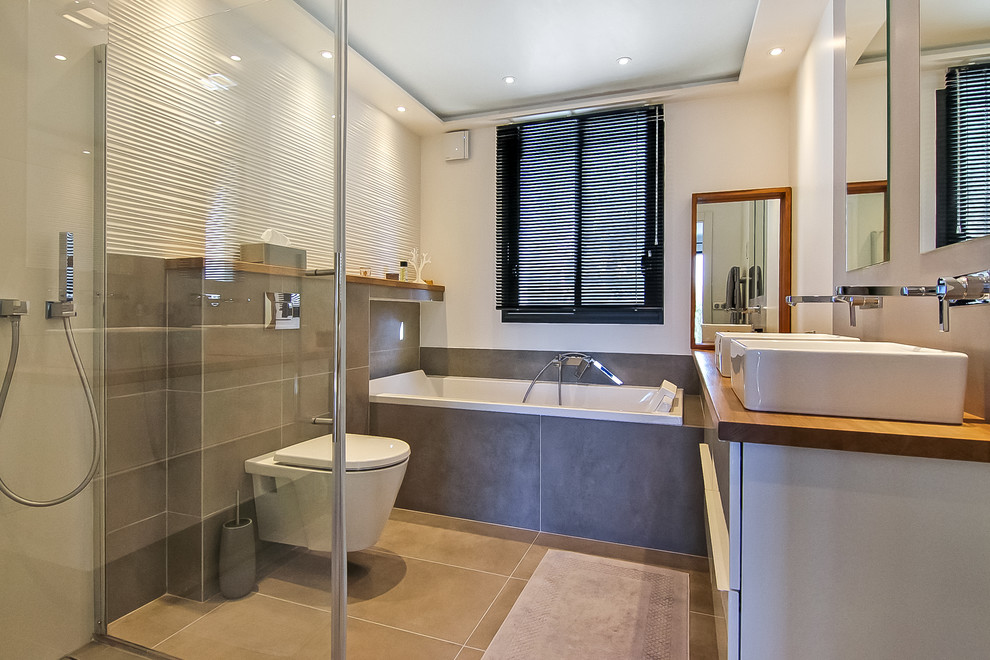 Photo of a mid-sized contemporary master bathroom in Nice with white cabinets, a drop-in tub, a curbless shower, a wall-mount toilet, white tile, ceramic tile, grey walls, ceramic floors, a drop-in sink, wood benchtops, grey floor, a hinged shower door and beige benchtops.