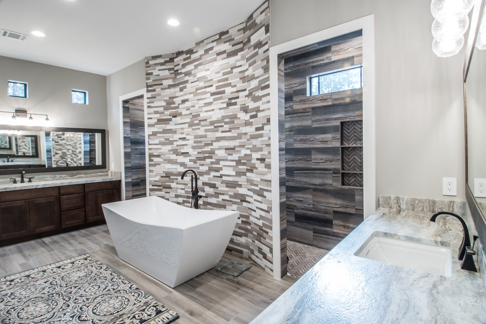 This is an example of a mid-sized contemporary master bathroom in Houston with recessed-panel cabinets, dark wood cabinets, a freestanding tub, an open shower, a two-piece toilet, brown tile, porcelain tile, grey walls, porcelain floors, an undermount sink, limestone benchtops, brown floor, an open shower and grey benchtops.