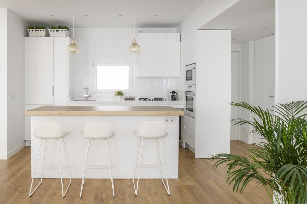 Inspiration for a contemporary l-shaped kitchen in Other with flat-panel cabinets, white cabinets, wood benchtops, white splashback, white appliances, with island, an undermount sink, glass sheet splashback, light hardwood floors, brown floor and beige benchtop.