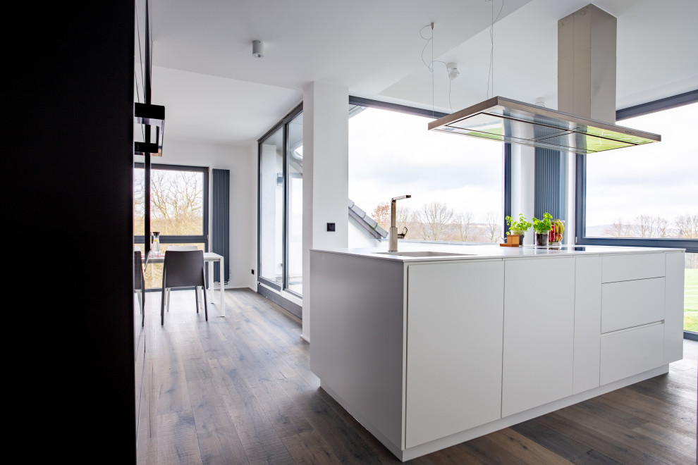 This is an example of a large modern open plan kitchen in Dortmund with an integrated sink, flat-panel cabinets, white cabinets, solid surface benchtops, black appliances, painted wood floors, with island and white benchtop.