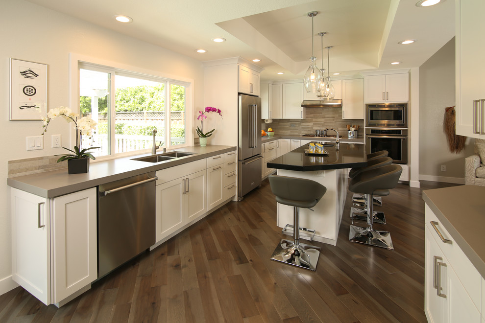 Photo of a large transitional l-shaped open plan kitchen in San Francisco with a double-bowl sink, shaker cabinets, white cabinets, beige splashback, matchstick tile splashback, stainless steel appliances, dark hardwood floors, with island, brown floor, quartz benchtops and grey benchtop.