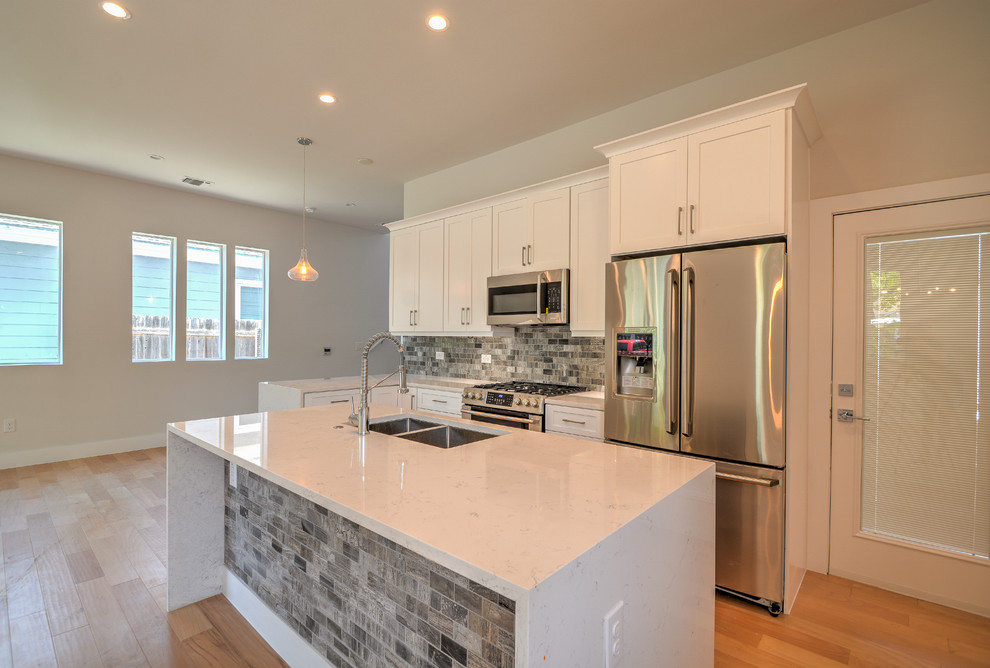 Inspiration for a mid-sized transitional single-wall medium tone wood floor and brown floor open concept kitchen remodel in Austin with a double-bowl sink, shaker cabinets, white cabinets, marble countertops, gray backsplash, stone tile backsplash, stainless steel appliances, an island and white countertops