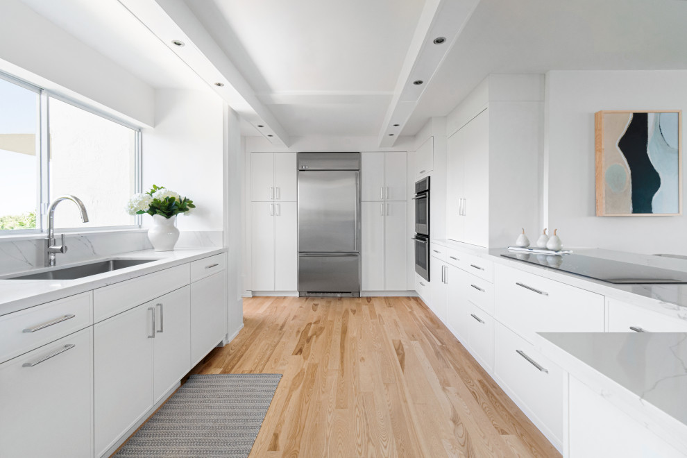 Design ideas for a beach style u-shaped kitchen in Miami with an undermount sink, flat-panel cabinets, white cabinets, white splashback, stainless steel appliances, light hardwood floors, a peninsula, beige floor and white benchtop.