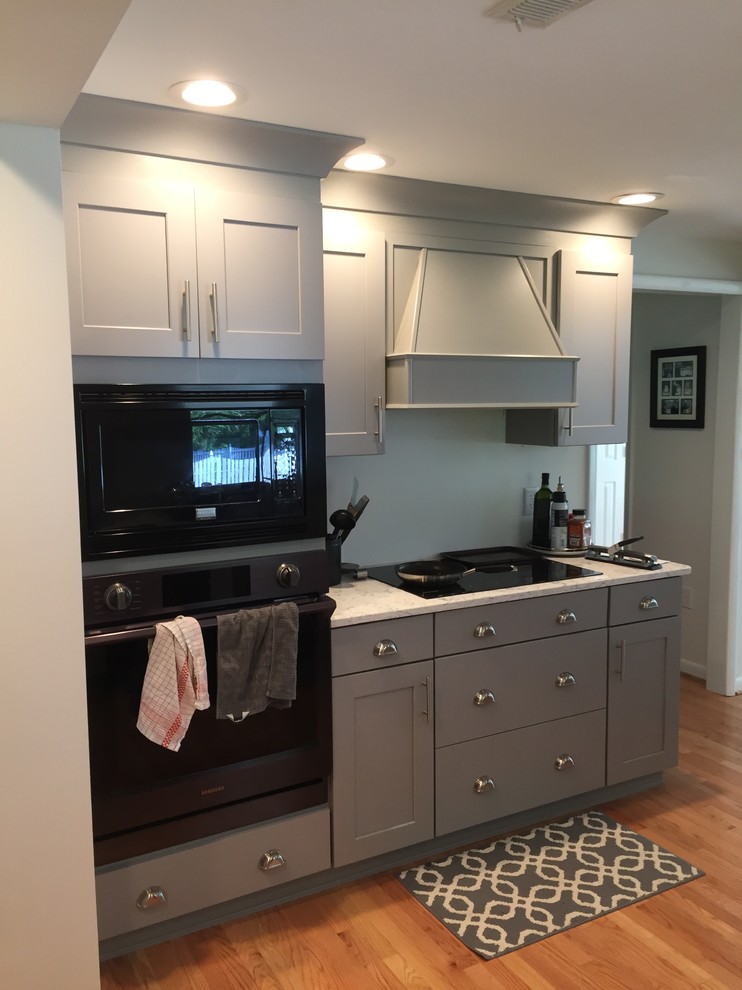 This is an example of a mid-sized traditional l-shaped open plan kitchen in DC Metro with a farmhouse sink, raised-panel cabinets, grey cabinets, granite benchtops, black appliances, medium hardwood floors, a peninsula and brown floor.