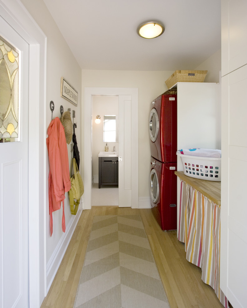 This is an example of a small transitional single-wall utility room in New York with a stacked washer and dryer, white cabinets, white walls, light hardwood floors and wood benchtops.