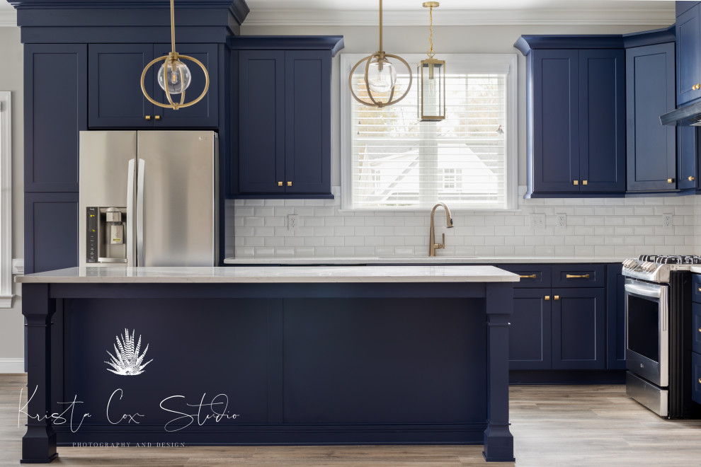 Example of a transitional light wood floor and brown floor kitchen design with an undermount sink, shaker cabinets, blue cabinets, quartz countertops, white backsplash, subway tile backsplash, stainless steel appliances, an island and white countertops