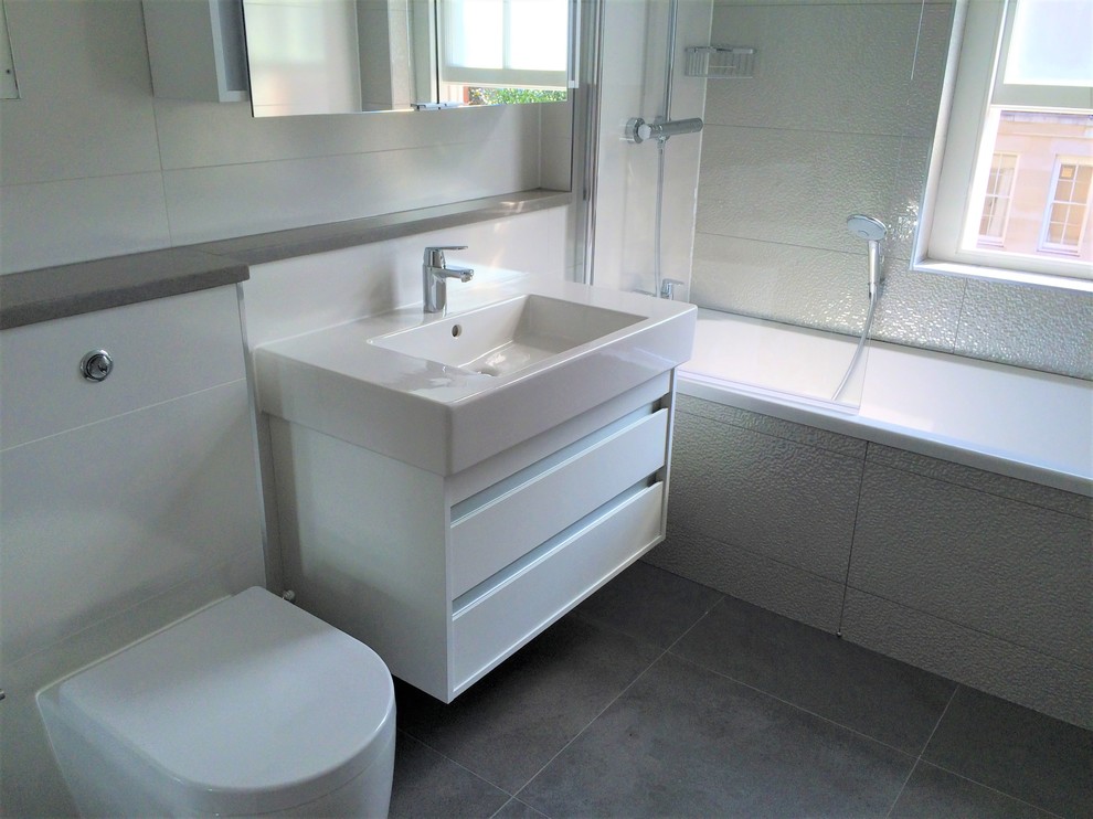 Photo of a small contemporary kids bathroom in London with flat-panel cabinets, white cabinets, a drop-in tub, a shower/bathtub combo, a one-piece toilet, white tile, ceramic tile, porcelain floors, a wall-mount sink, quartzite benchtops, grey floor, a hinged shower door and grey benchtops.