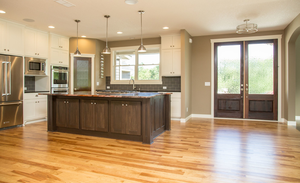 Example of a transitional l-shaped light wood floor eat-in kitchen design in Minneapolis with a farmhouse sink, shaker cabinets, white cabinets, granite countertops, gray backsplash, ceramic backsplash, stainless steel appliances and an island