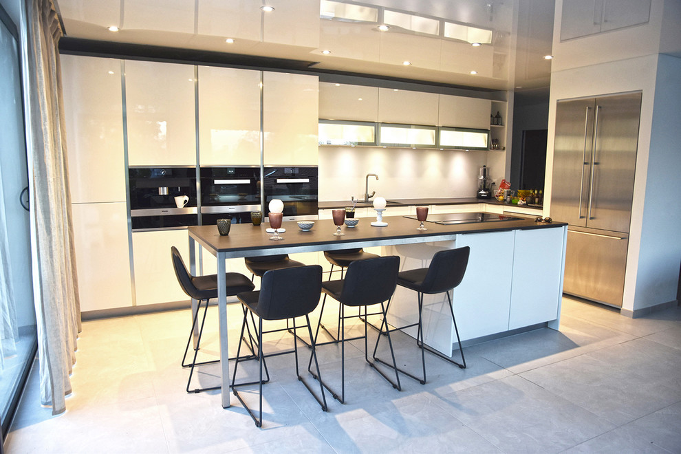 Large modern l-shaped open plan kitchen with an undermount sink, white cabinets, white splashback, with island, grey floor, beaded inset cabinets, limestone benchtops, glass sheet splashback, black appliances and ceramic floors.