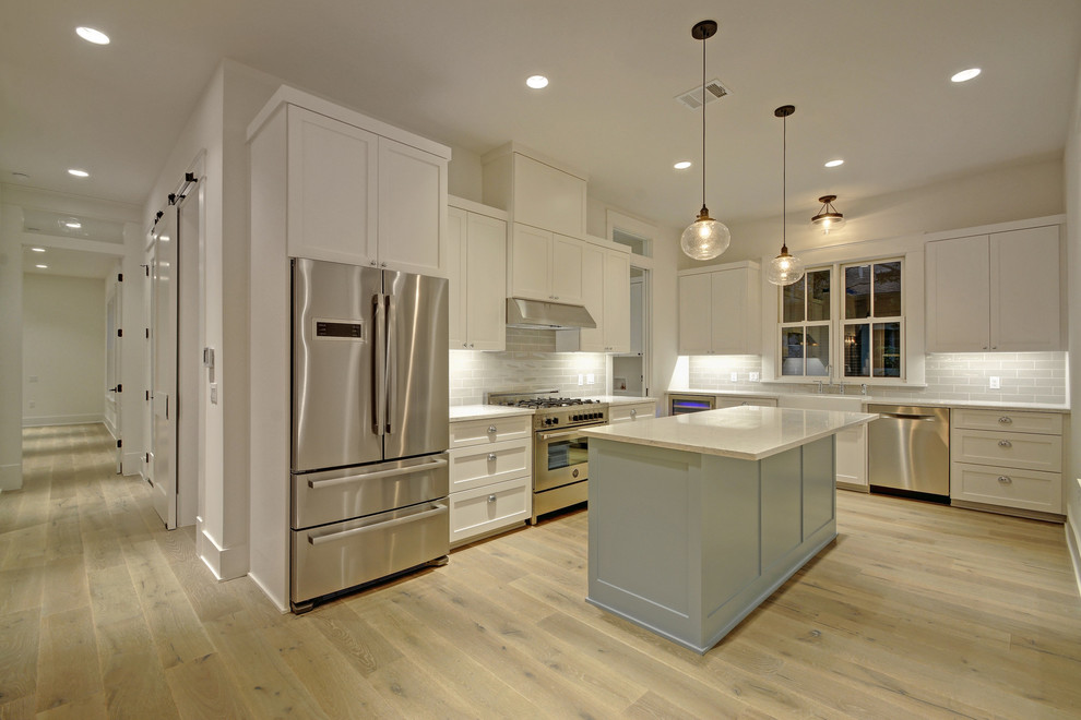 Mid-sized country l-shaped open plan kitchen in Austin with a farmhouse sink, shaker cabinets, white cabinets, ceramic splashback, stainless steel appliances, laminate floors, with island and beige floor.