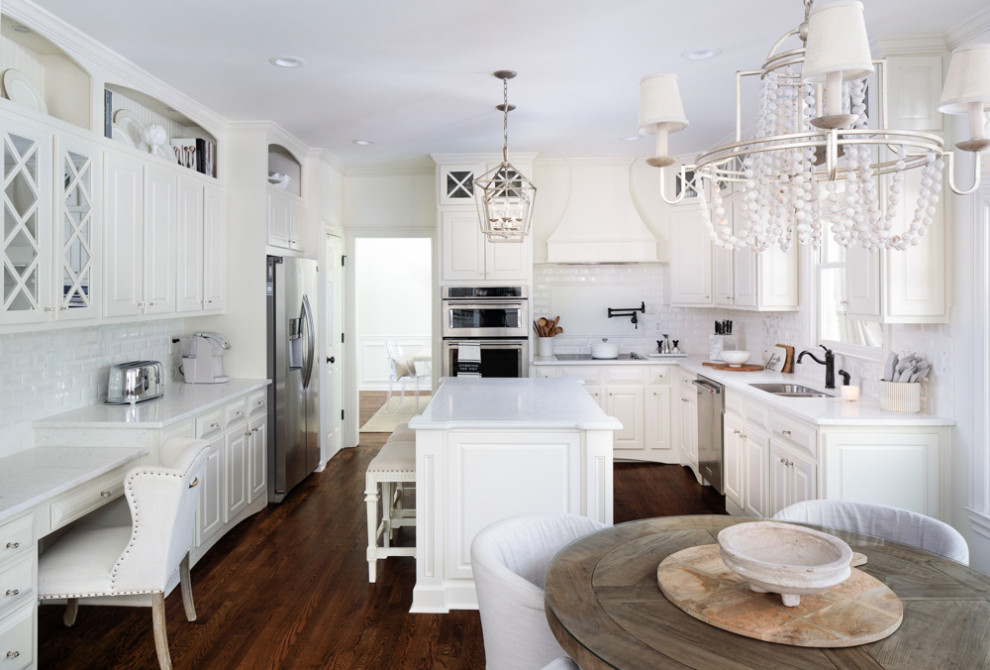 Shabby chic-inspirerad inredning av ett stort vit vitt kök, med en undermonterad diskho, luckor med upphöjd panel, vita skåp, bänkskiva i kvarts, vitt stänkskydd, stänkskydd i keramik, rostfria vitvaror, mörkt trägolv, en köksö och brunt golv
