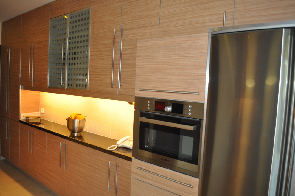 Mid-sized minimalist galley ceramic tile and beige floor enclosed kitchen photo with a double-bowl sink, flat-panel cabinets, medium tone wood cabinets, granite countertops, beige backsplash, ceramic backsplash, stainless steel appliances, an island and black countertops