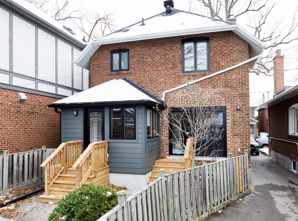 This is an example of a mid-sized transitional two-storey brick red house exterior in Toronto with a clipped gable roof, a shingle roof, a black roof and clapboard siding.