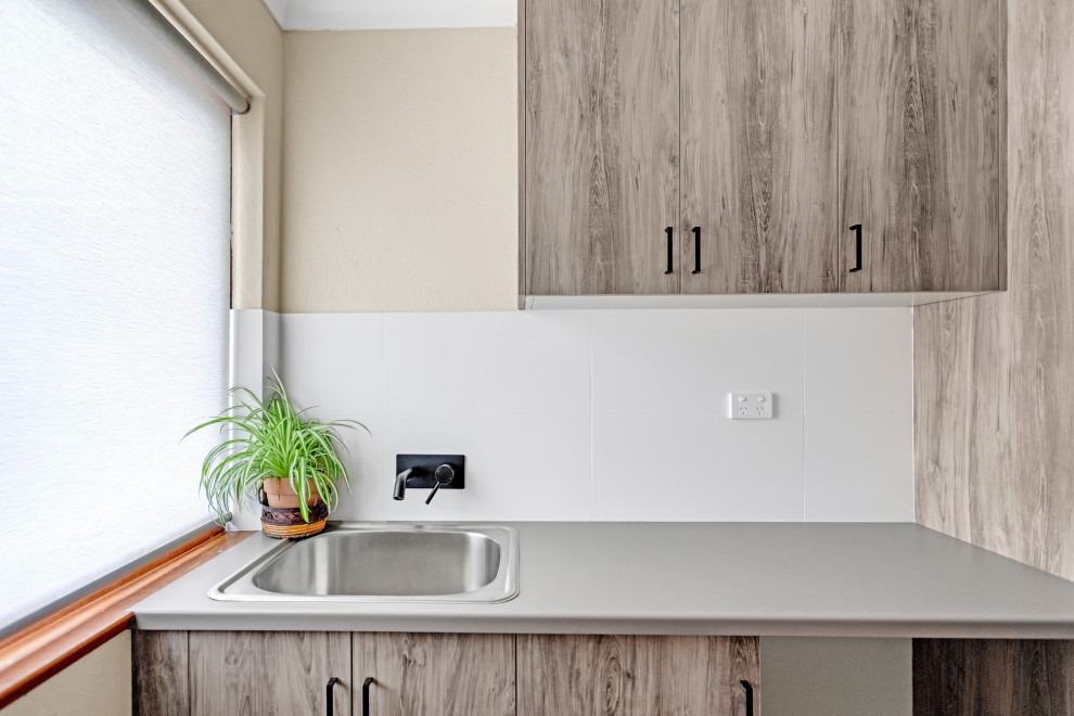Photo of a small transitional single-wall dedicated laundry room in Adelaide with a drop-in sink, flat-panel cabinets, light wood cabinets, laminate benchtops, white splashback, white walls, porcelain floors, a stacked washer and dryer, grey floor and grey benchtop.