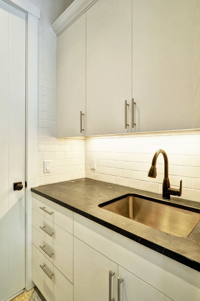 Design ideas for a mid-sized traditional galley dedicated laundry room in New York with an undermount sink, flat-panel cabinets, white cabinets, soapstone benchtops, white walls, travertine floors and a stacked washer and dryer.