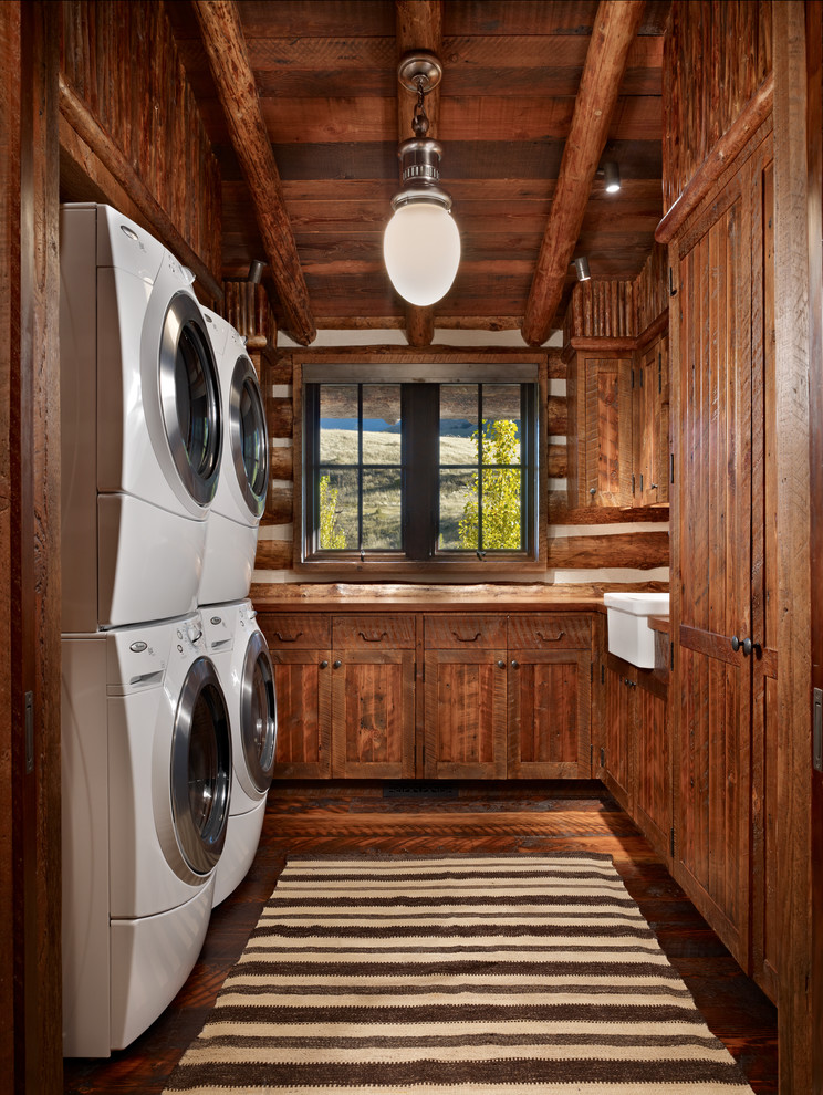 Photo of a mid-sized country dedicated laundry room in Other with a farmhouse sink, shaker cabinets, dark wood cabinets, wood benchtops, brown walls, dark hardwood floors, a stacked washer and dryer, brown floor and brown benchtop.