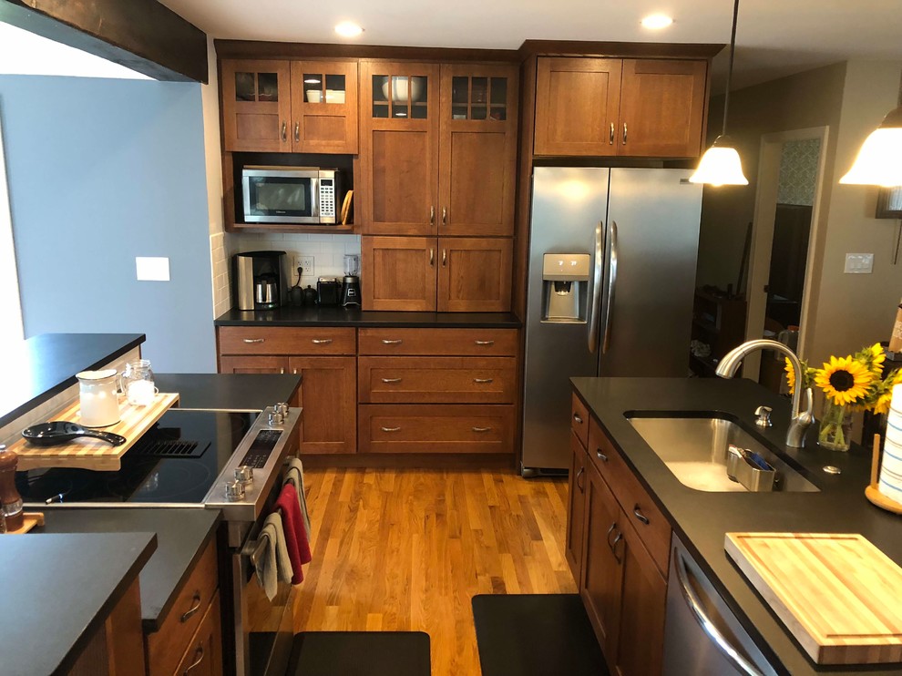 Design ideas for an arts and crafts galley eat-in kitchen in Portland Maine with an undermount sink, shaker cabinets, granite benchtops, white splashback, subway tile splashback, stainless steel appliances, light hardwood floors, multiple islands, beige floor, black benchtop and medium wood cabinets.