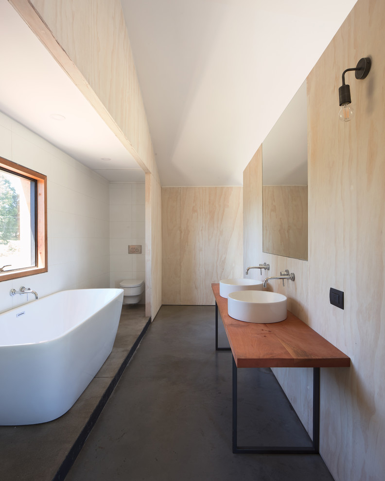 Inspiration for a large modern bathroom in Other with a freestanding tub, a wall-mount toilet, white tile, ceramic tile, white walls, concrete floors, a vessel sink, wood benchtops, grey floor and brown benchtops.