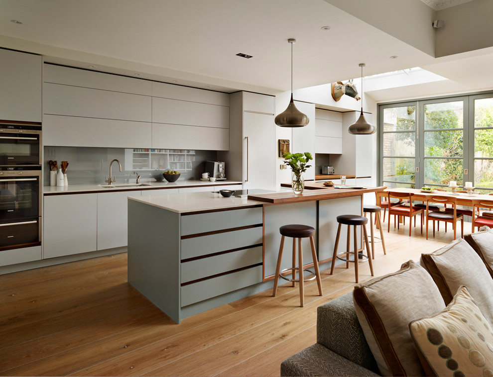 Inspiration for a large contemporary galley open plan kitchen in London with flat-panel cabinets, solid surface benchtops, grey splashback, stainless steel appliances, with island, white benchtop, an undermount sink, grey cabinets, glass sheet splashback, medium hardwood floors and brown floor.