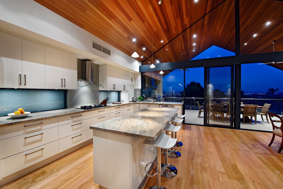 Photo of a mid-sized modern galley eat-in kitchen in Perth with blue splashback, a drop-in sink, white cabinets, granite benchtops, glass sheet splashback, stainless steel appliances, medium hardwood floors and with island.