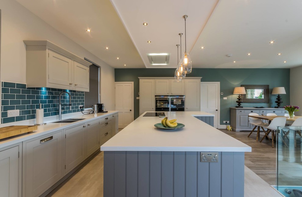 Photo of a large contemporary open plan kitchen in Cheshire with a drop-in sink, recessed-panel cabinets, white cabinets, quartz benchtops, blue splashback, subway tile splashback, panelled appliances, light hardwood floors and with island.