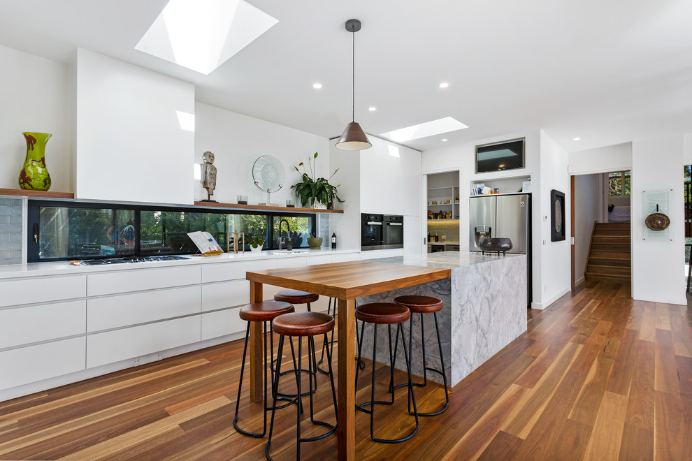 Design ideas for a mid-sized contemporary galley open plan kitchen in Melbourne with a drop-in sink, flat-panel cabinets, white cabinets, marble benchtops, glass sheet splashback, stainless steel appliances, light hardwood floors, with island, brown floor and grey benchtop.