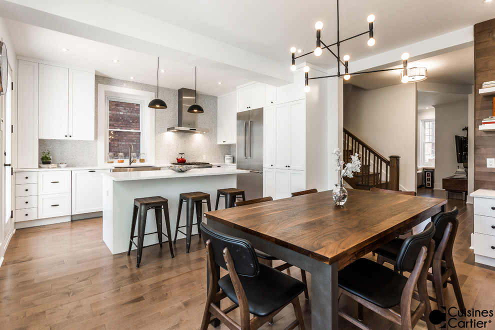 Example of a mid-sized transitional l-shaped medium tone wood floor eat-in kitchen design in Montreal with a farmhouse sink, shaker cabinets, white cabinets, quartzite countertops, white backsplash, mosaic tile backsplash, stainless steel appliances and an island