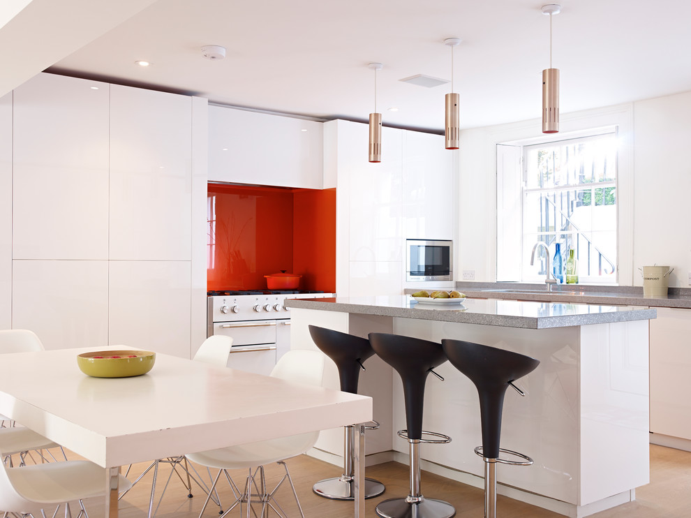 Large contemporary eat-in kitchen in London with with island, flat-panel cabinets, white cabinets, solid surface benchtops, orange splashback, glass tile splashback and white appliances.
