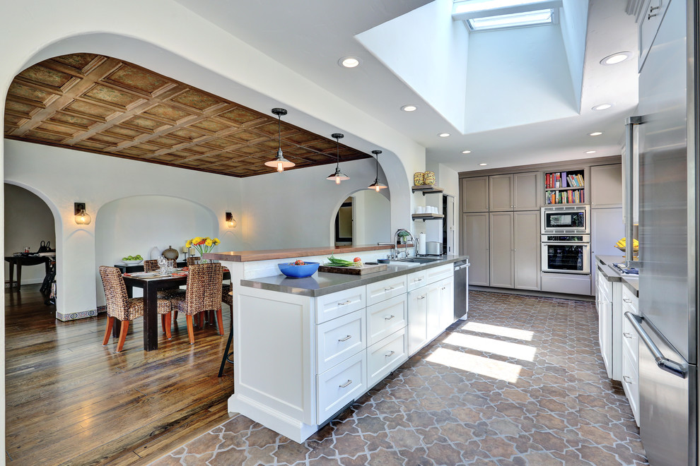 This is an example of a large mediterranean l-shaped eat-in kitchen in Los Angeles with shaker cabinets, white cabinets, stainless steel appliances, solid surface benchtops, with island, an undermount sink, beige splashback, brick splashback, terra-cotta floors and multi-coloured floor.