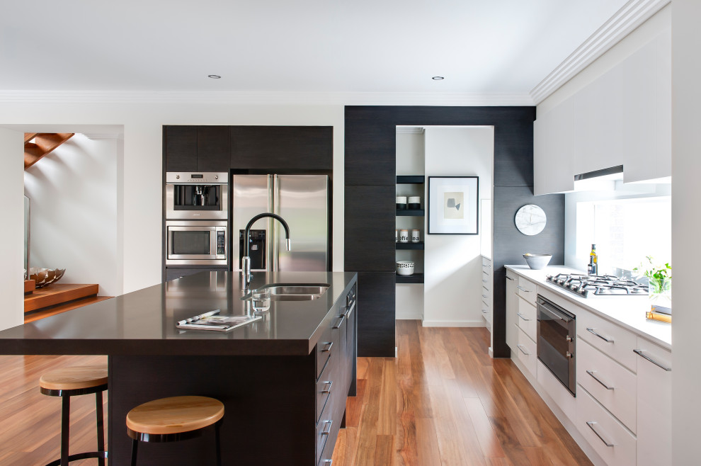 This is an example of a contemporary l-shaped open plan kitchen in Sydney with stainless steel appliances, with island, a double-bowl sink, flat-panel cabinets, medium hardwood floors, brown floor and white benchtop.