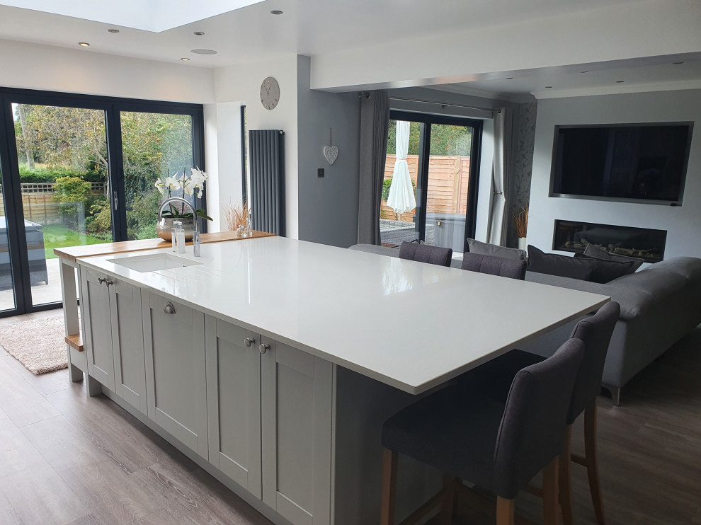 This is an example of a large contemporary l-shaped open plan kitchen in West Midlands with an integrated sink, shaker cabinets, grey cabinets, quartzite benchtops, white splashback, engineered quartz splashback, black appliances, laminate floors, with island, grey floor and white benchtop.