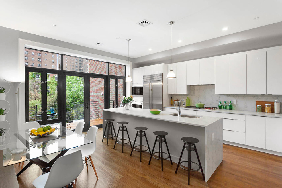 Photo of a large modern single-wall eat-in kitchen in New York with an undermount sink, flat-panel cabinets, white cabinets, marble benchtops, white splashback, stone slab splashback, stainless steel appliances, medium hardwood floors, with island and brown floor.