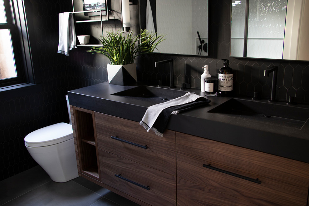 This is an example of a small modern kids bathroom in Salt Lake City with flat-panel cabinets, medium wood cabinets, a corner tub, a shower/bathtub combo, a one-piece toilet, black tile, ceramic tile, black walls, ceramic floors, an integrated sink, engineered quartz benchtops, grey floor, a hinged shower door, black benchtops, a double vanity and a floating vanity.