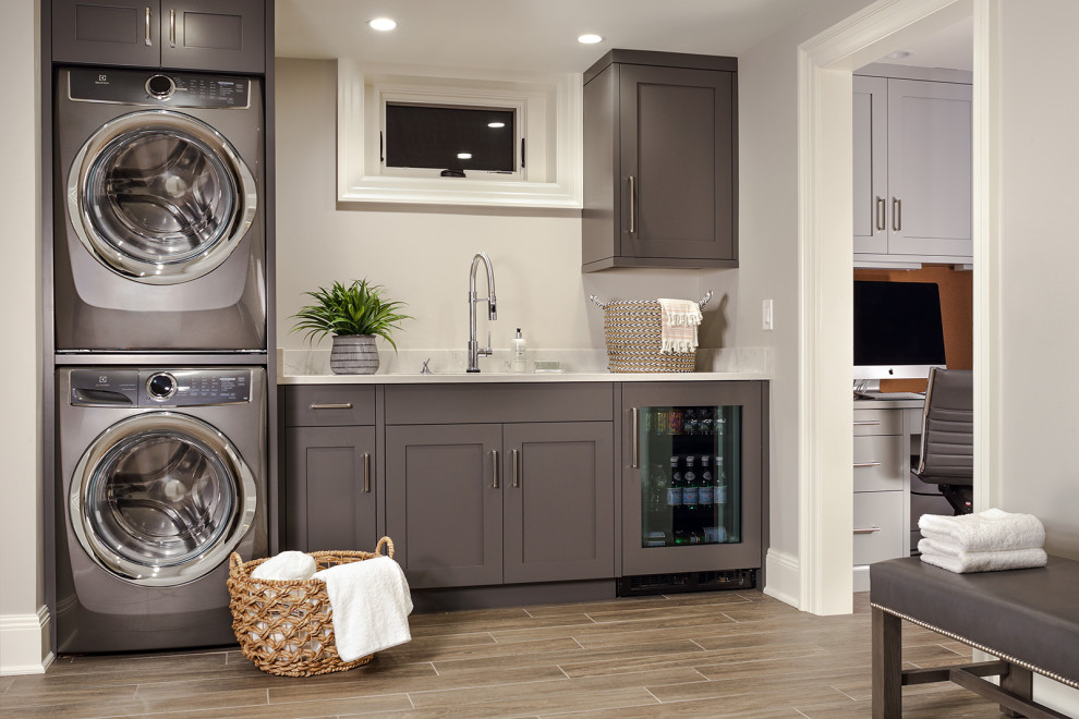 Design ideas for a mid-sized transitional single-wall utility room in New York with an undermount sink, flat-panel cabinets, grey cabinets, quartz benchtops, grey walls, ceramic floors, a stacked washer and dryer, brown floor and white benchtop.
