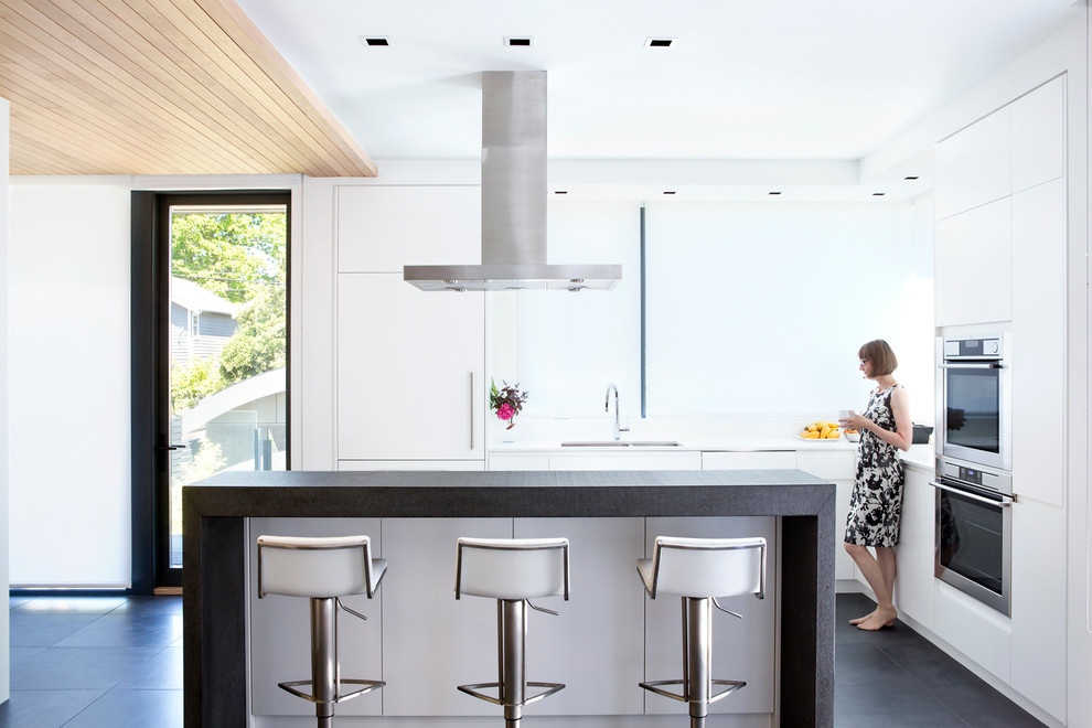 Photo of a small contemporary kitchen in Vancouver with a double-bowl sink, flat-panel cabinets, white cabinets, quartz benchtops, white splashback, stainless steel appliances, slate floors and with island.