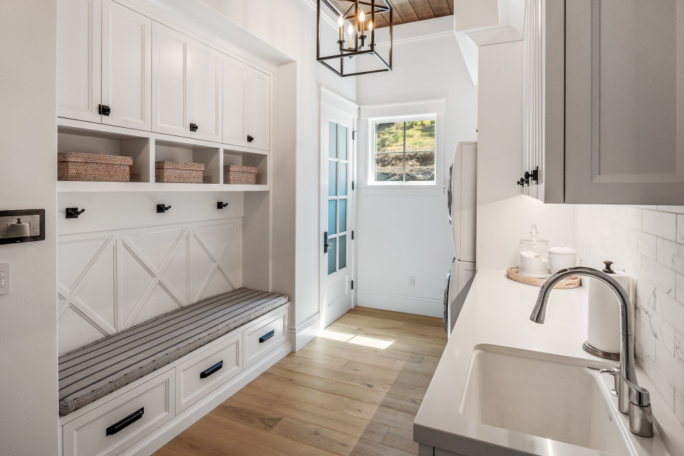 Design ideas for a large transitional galley dedicated laundry room in San Francisco with an undermount sink, shaker cabinets, white cabinets, white splashback, ceramic splashback, white walls, light hardwood floors, a stacked washer and dryer, beige floor, white benchtop and exposed beam.