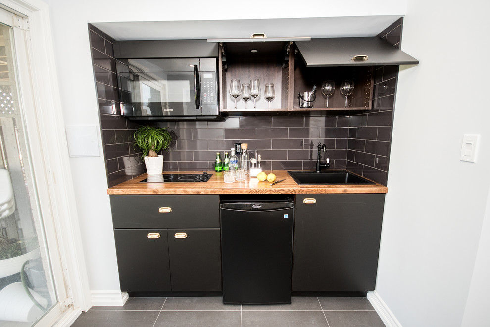 Small contemporary single-wall kitchen pantry in Toronto with a drop-in sink, flat-panel cabinets, black cabinets, wood benchtops, black splashback, ceramic splashback, black appliances, porcelain floors, no island, grey floor and brown benchtop.
