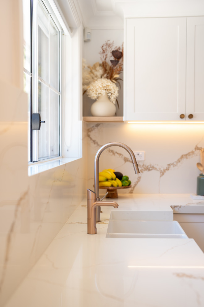 Photo of a mid-sized u-shaped kitchen pantry in Sydney with a farmhouse sink, recessed-panel cabinets, white cabinets, quartz benchtops, white splashback, engineered quartz splashback, white appliances, laminate floors, with island, brown floor and white benchtop.