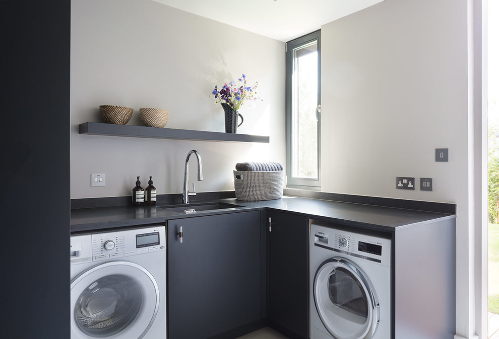 This is an example of a large contemporary single-wall laundry room in London with an undermount sink, marble benchtops, light hardwood floors and grey cabinets.
