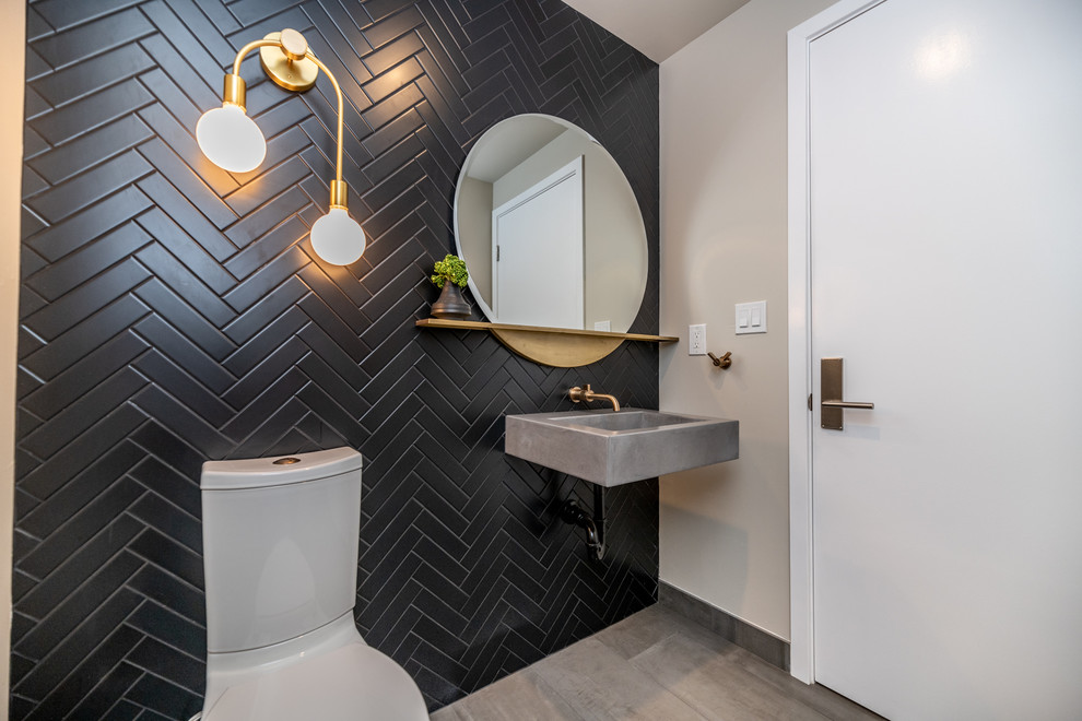 Photo of a small midcentury powder room in Los Angeles with a two-piece toilet, black tile, ceramic tile, black walls, porcelain floors, an integrated sink, concrete benchtops, grey floor and grey benchtops.