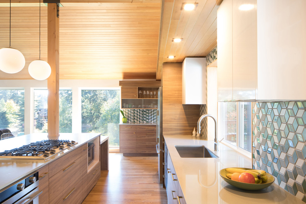 This is an example of a mid-sized midcentury u-shaped open plan kitchen in Seattle with a single-bowl sink, flat-panel cabinets, dark wood cabinets, quartz benchtops, mosaic tile splashback, stainless steel appliances, medium hardwood floors, with island, brown floor and white benchtop.
