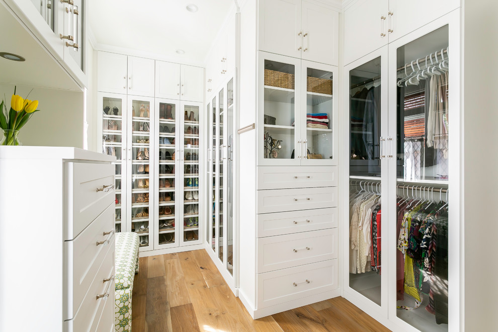 This is an example of a gender-neutral walk-in wardrobe in Charleston with glass-front cabinets, white cabinets, medium hardwood floors and brown floor.