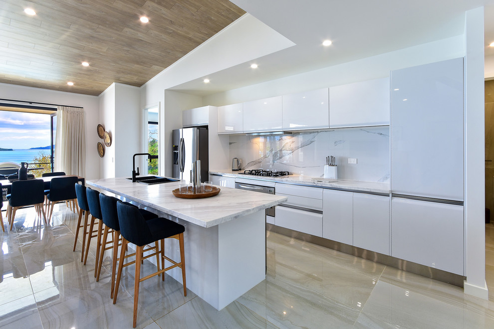 Inspiration for a contemporary galley eat-in kitchen in Brisbane with a double-bowl sink, flat-panel cabinets, white cabinets, white splashback, stainless steel appliances, with island, beige floor and white benchtop.
